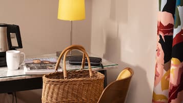 A hotel room with a glass-top desk, a wicker basket, a white mug, a magazine, a lamp, and a colorful curtain.