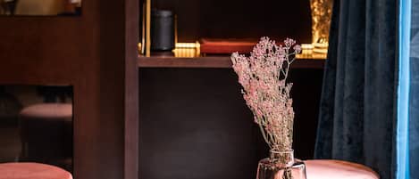Una mesa redonda de cristal con un florero de flores secas, dos otomanas rosadas y una estantería de madera oscura con libros y objetos decorativos.