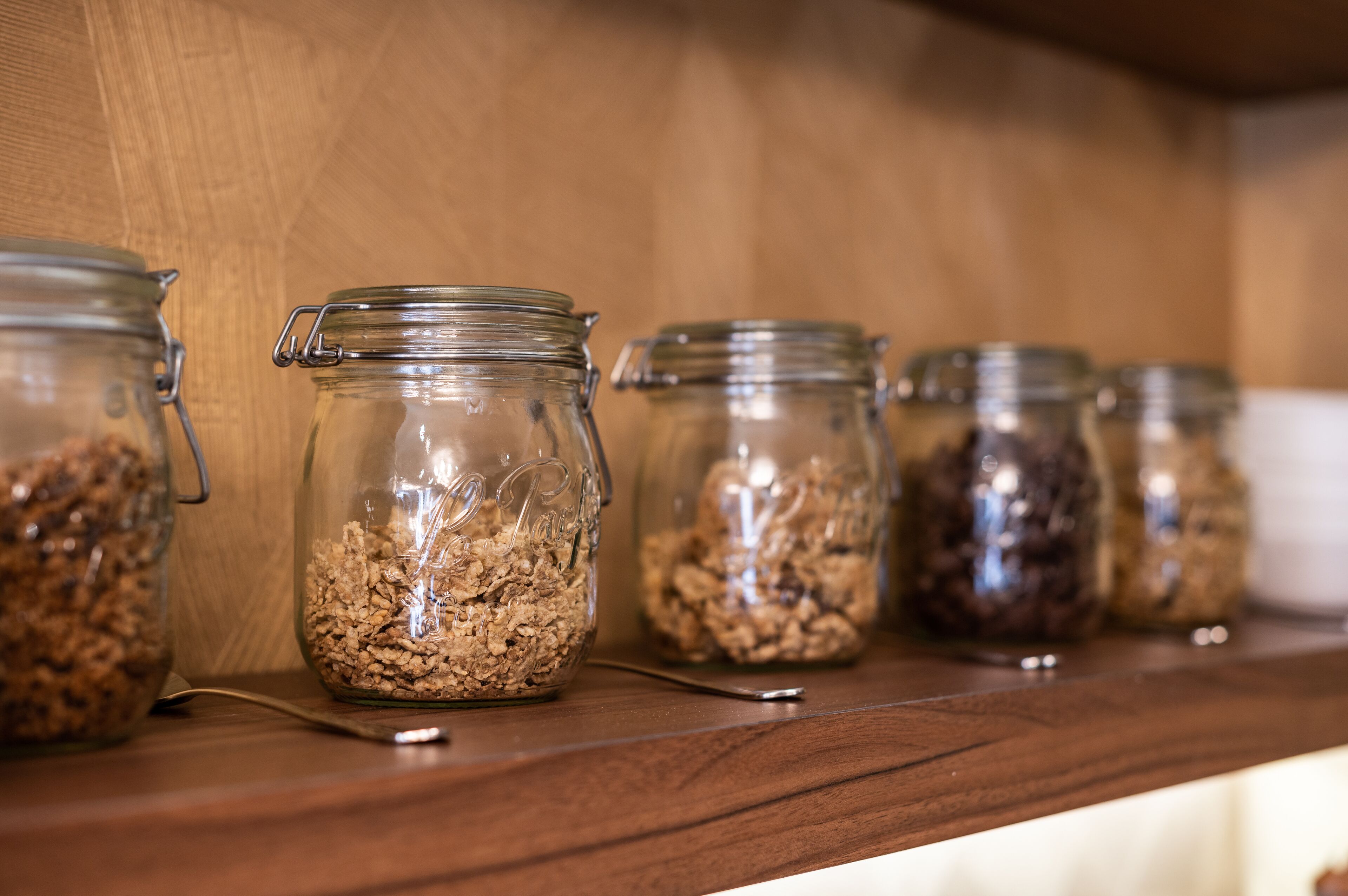 Une rangée de bocaux en verre contenant du granola, posés sur une étagère en bois.