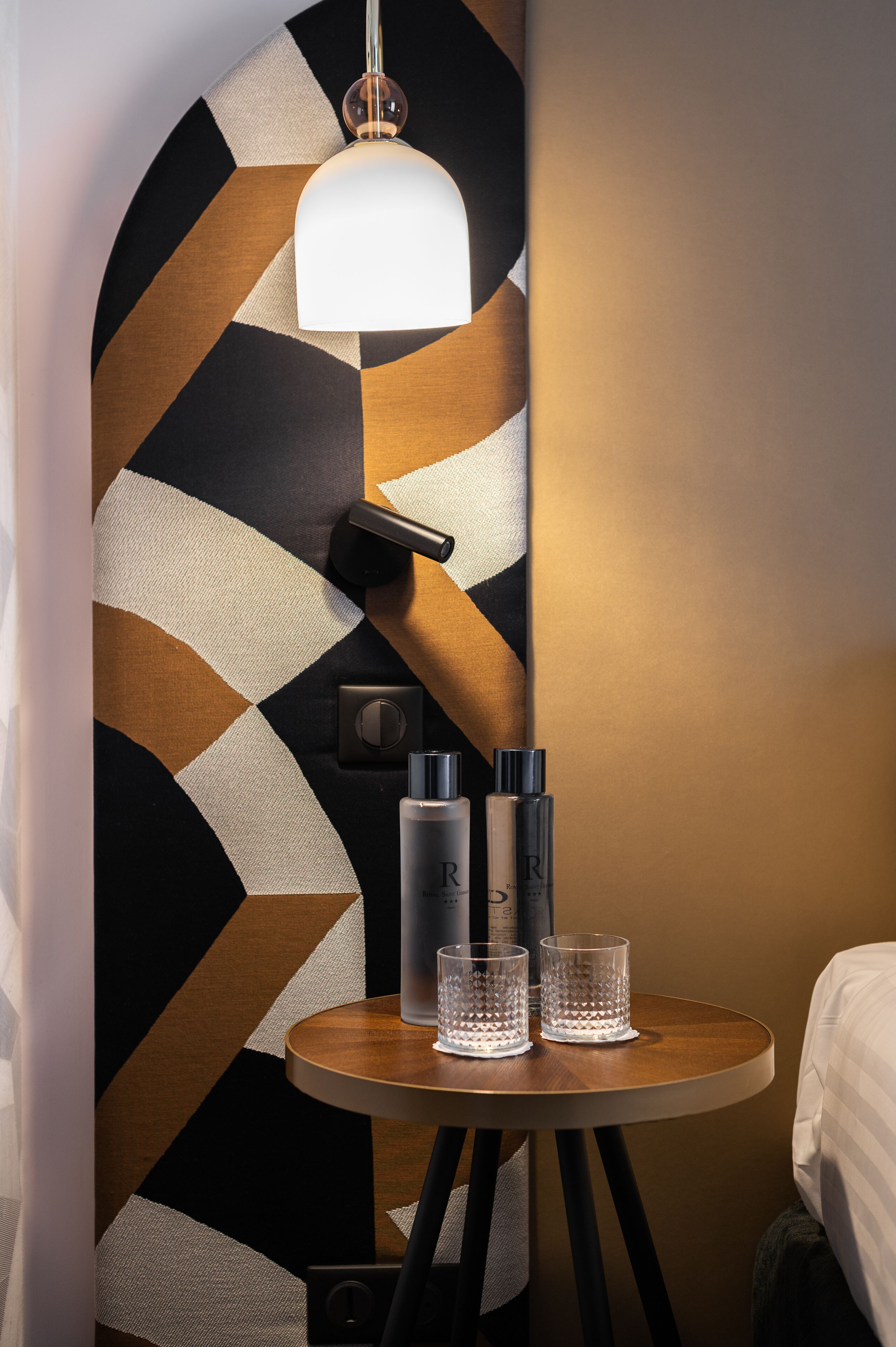 A small round table with two glasses and two bottles, a wall with a geometric pattern, and a hanging light fixture.