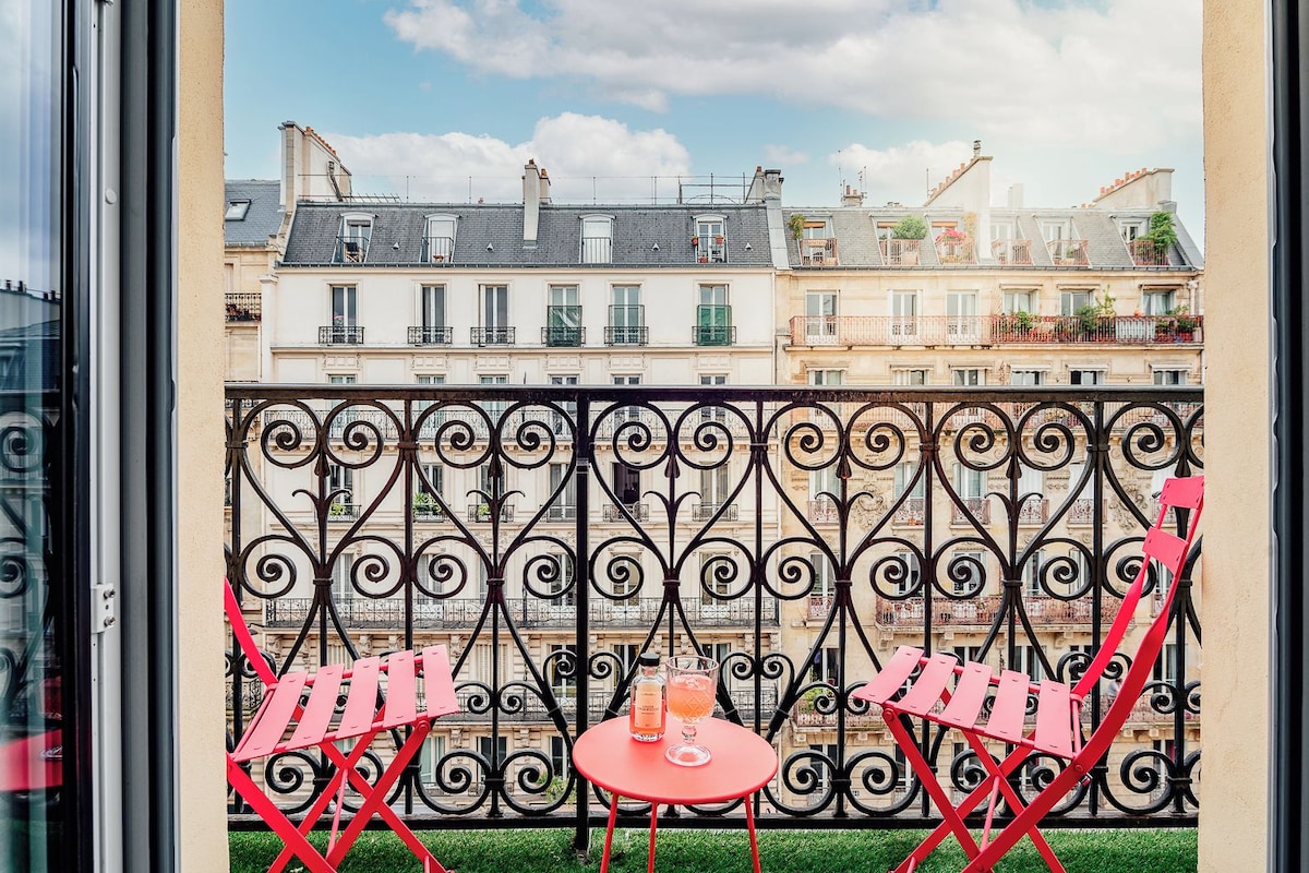 A balcony with wrought iron railings, two red chairs, and a small table with a drink.