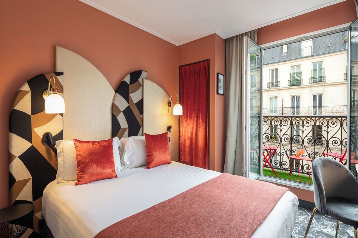 Habitación de hotel con una cama grande, dos lámparas fijadas a la pared, una cortina roja y un balcón con vistas a edificios.