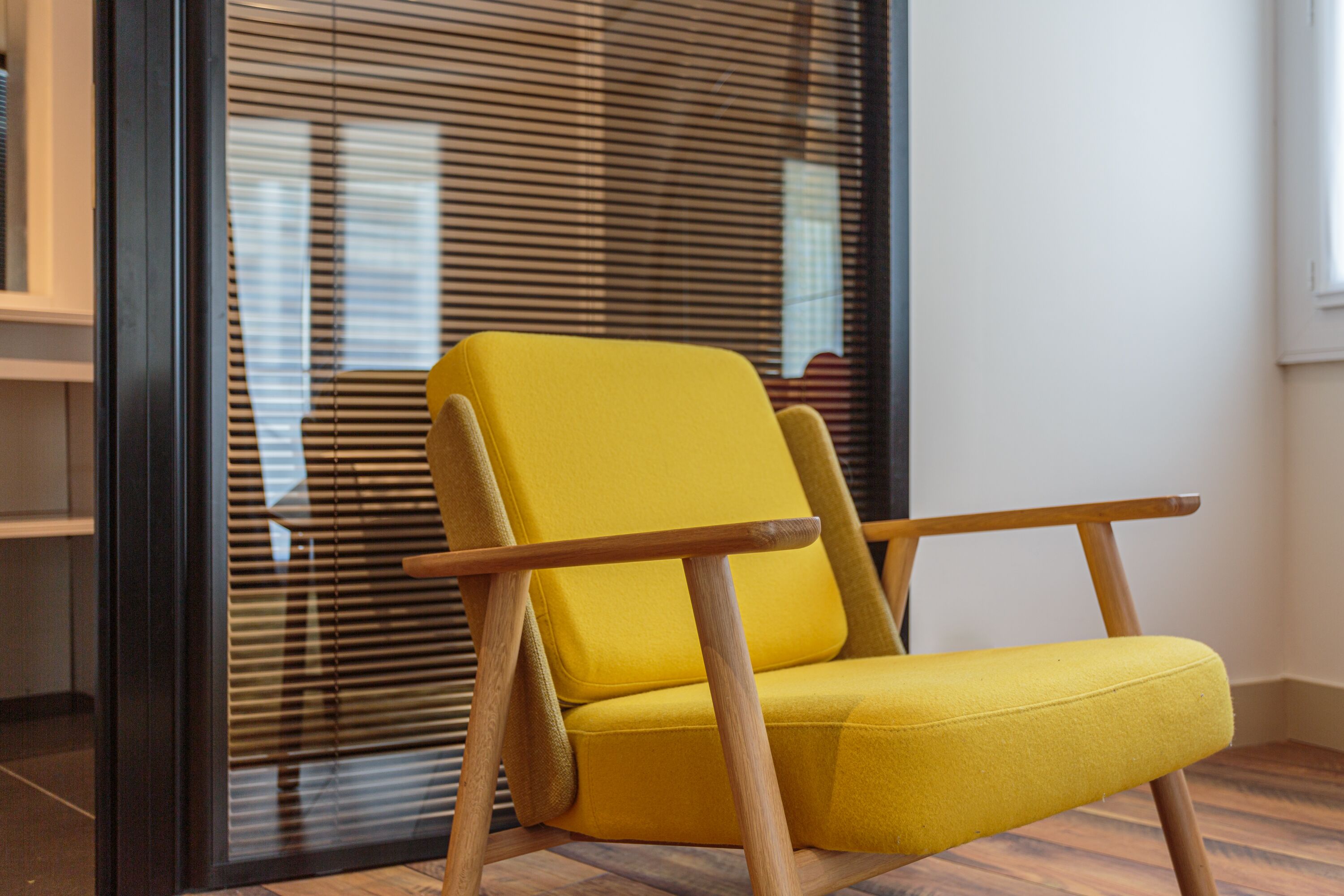 Un sillón amarillo con patas y reposabrazos de madera en una habitación con piso de madera y una ventana con persianas.