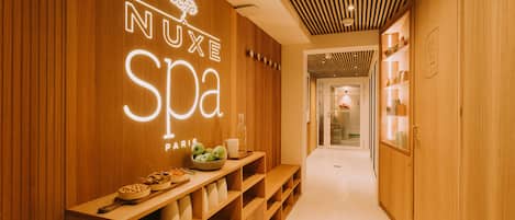 A wooden interior hallway with a sign reading "NUXE SPA PARIS" and a reception desk with bowls of fruit and cups.