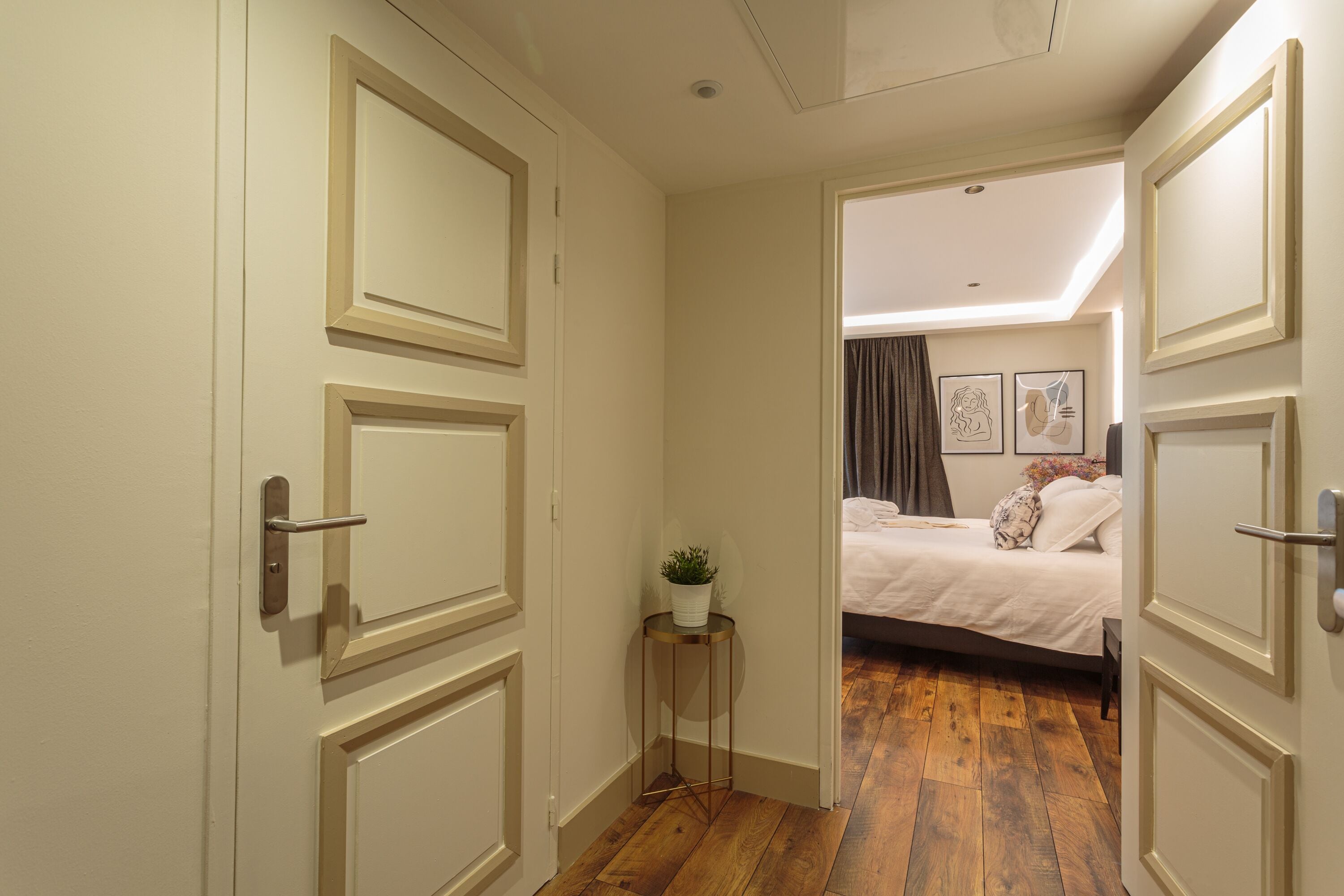 Un estrecho pasillo con suelo de madera que lleva a un dormitorio con cama y una pequeña mesa con una planta.