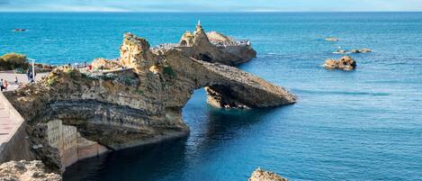 A coastal rock formation with a bridge, clear blue sea, and rocky terrain.