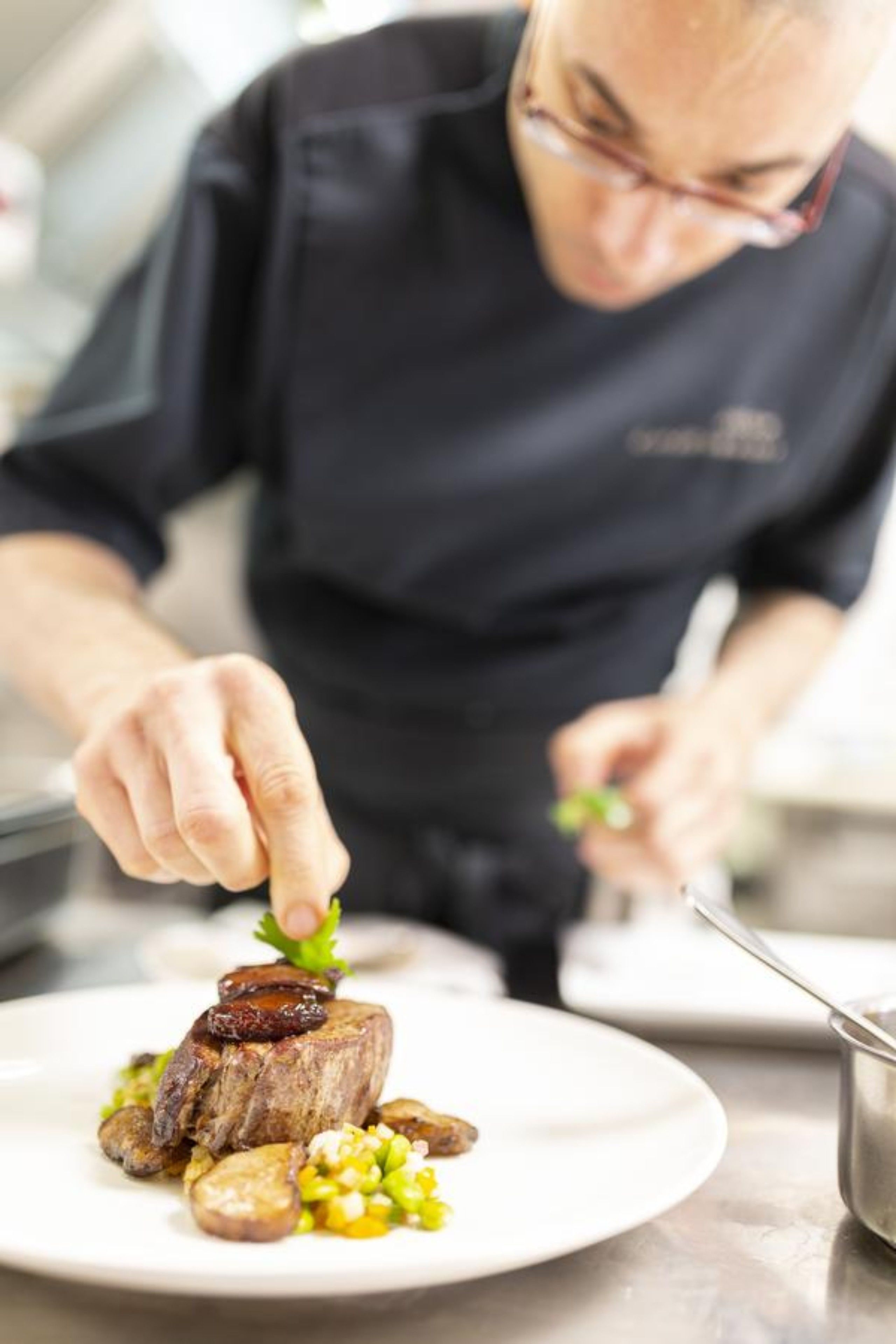 En kok tilbereder en ret med steak, kartofler og majs på en hvid tallerken.