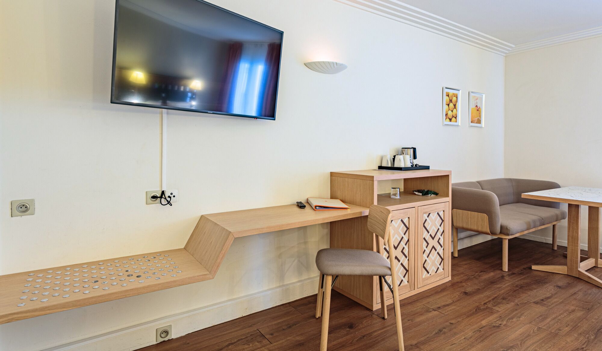 Ein modernes Wohnzimmer mit Flachbildfernseher an der Wand, einer Holzbank mit Stauraum, einer kleinen Kommode mit Zierelementen, einem Sofa und einem Esstisch.