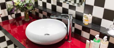 Un baño con lavamanos blanco, grifo cromado y encimera roja. Hay un espejo sobre el lavamanos y un salpicadero con baldosas de blanco y negro a cuadros.
