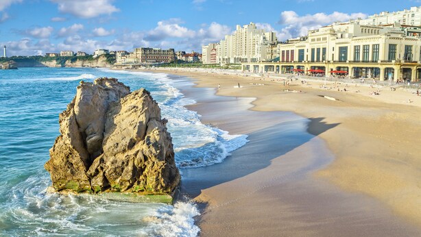 A coastal beach with a prominent rock formation, a sandy shore, and a row of buildings with large windows and balconies. - Grand Tonic Hotel & SPA NUXE (Biarritz)