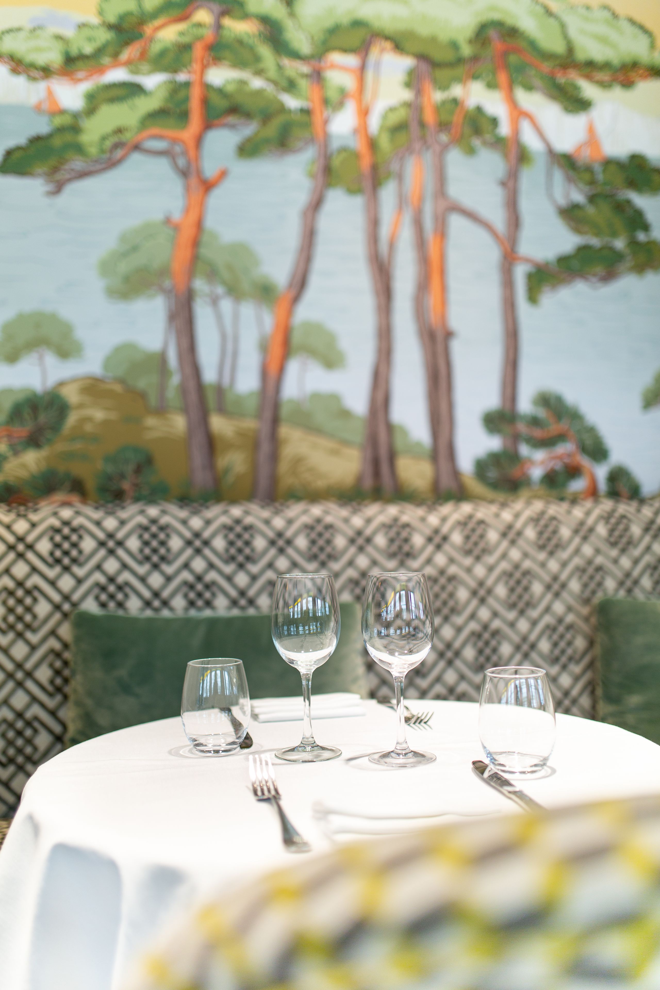 Una mesa de comedor con tres copas de vino y cubiertos, frente a un mural que representa árboles y follaje.