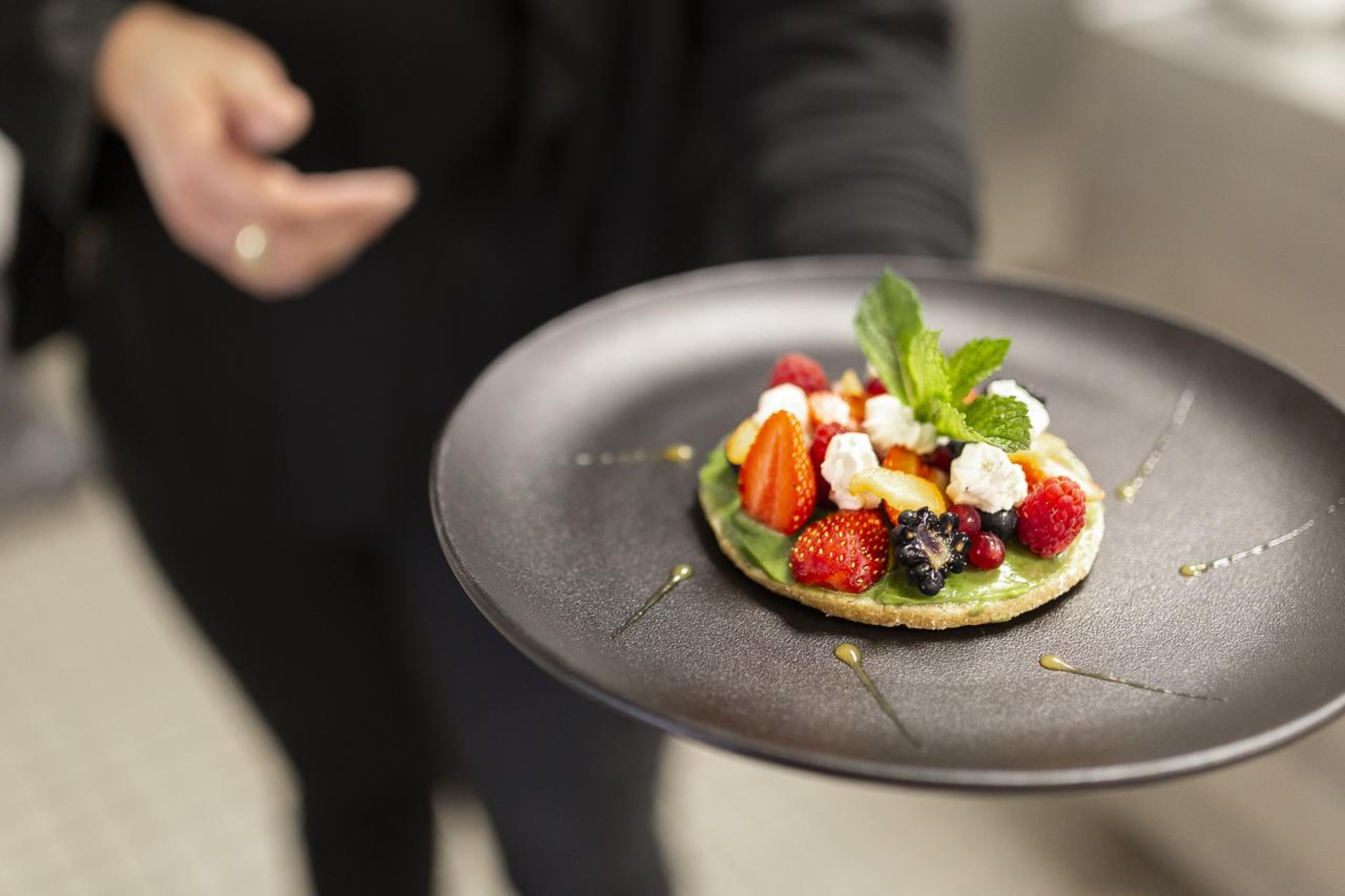 A person holding a plate with a dessert featuring strawberries, raspberries, and a mint leaf.