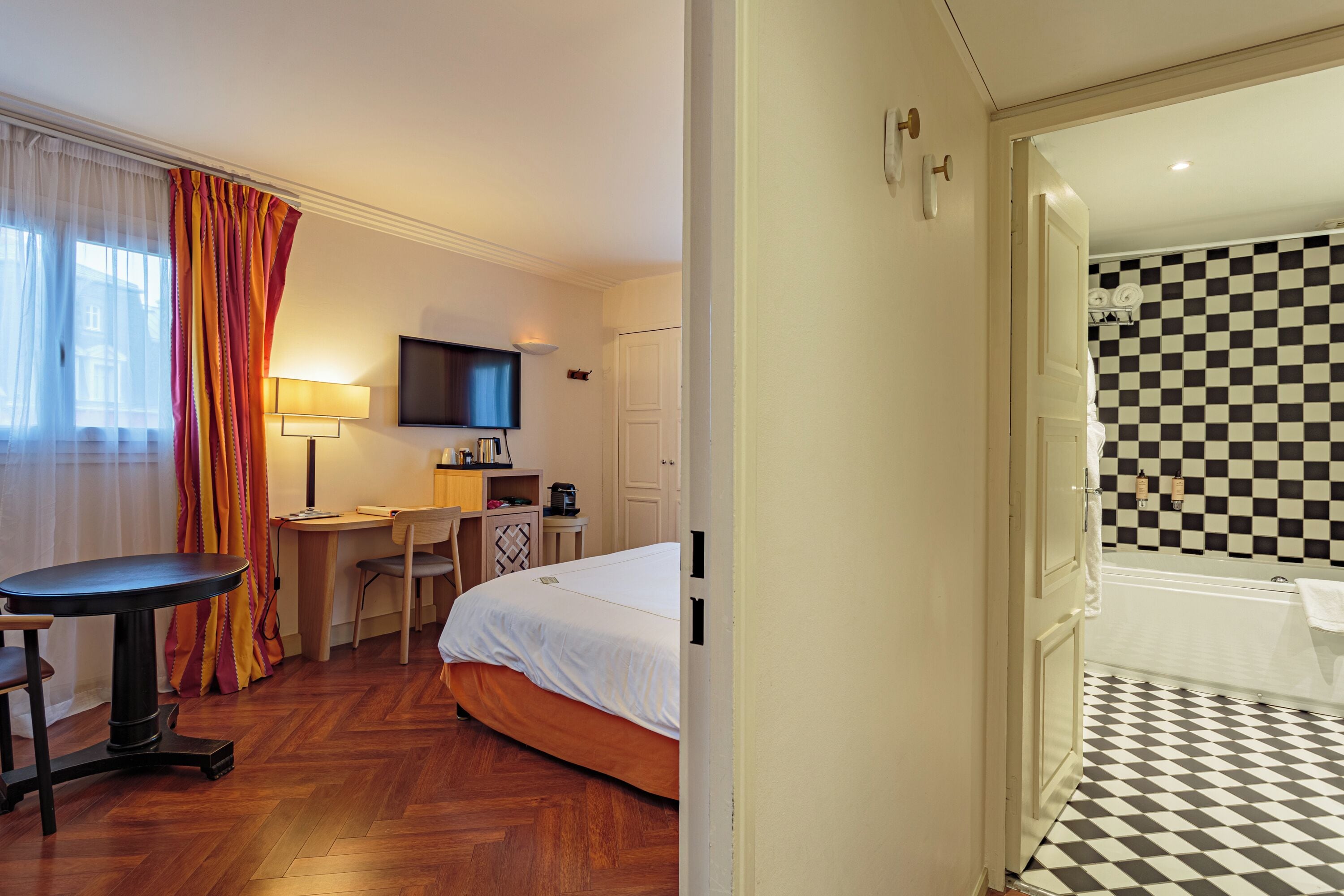 A hotel room with a bed, a desk, a chair, a TV, and a bathroom with a checkered floor.