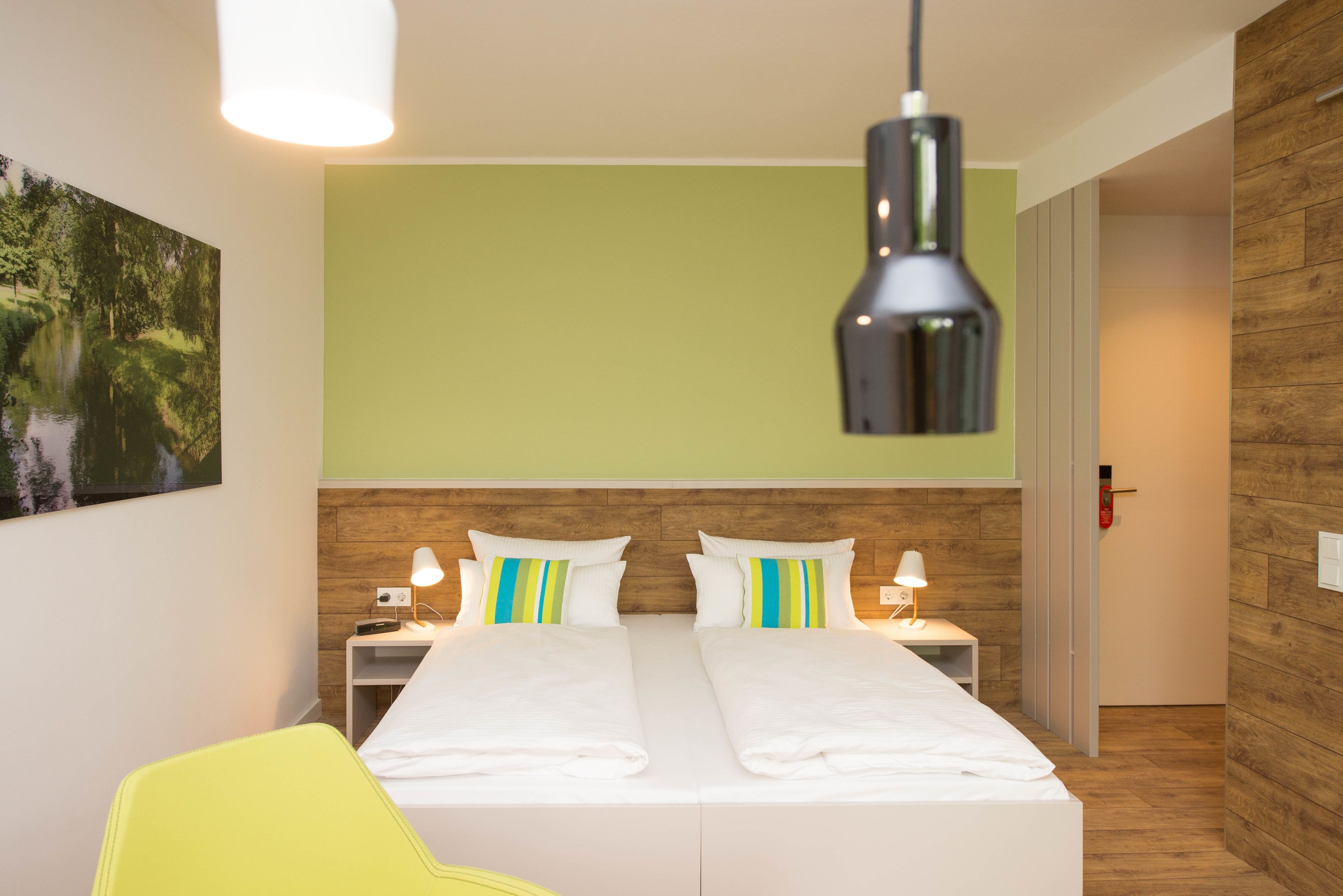 Een tweepersoonsbed met wit beddengoed en gestreepte kussens, een nachtkastje met een lamp, en een houten hoofdbord.