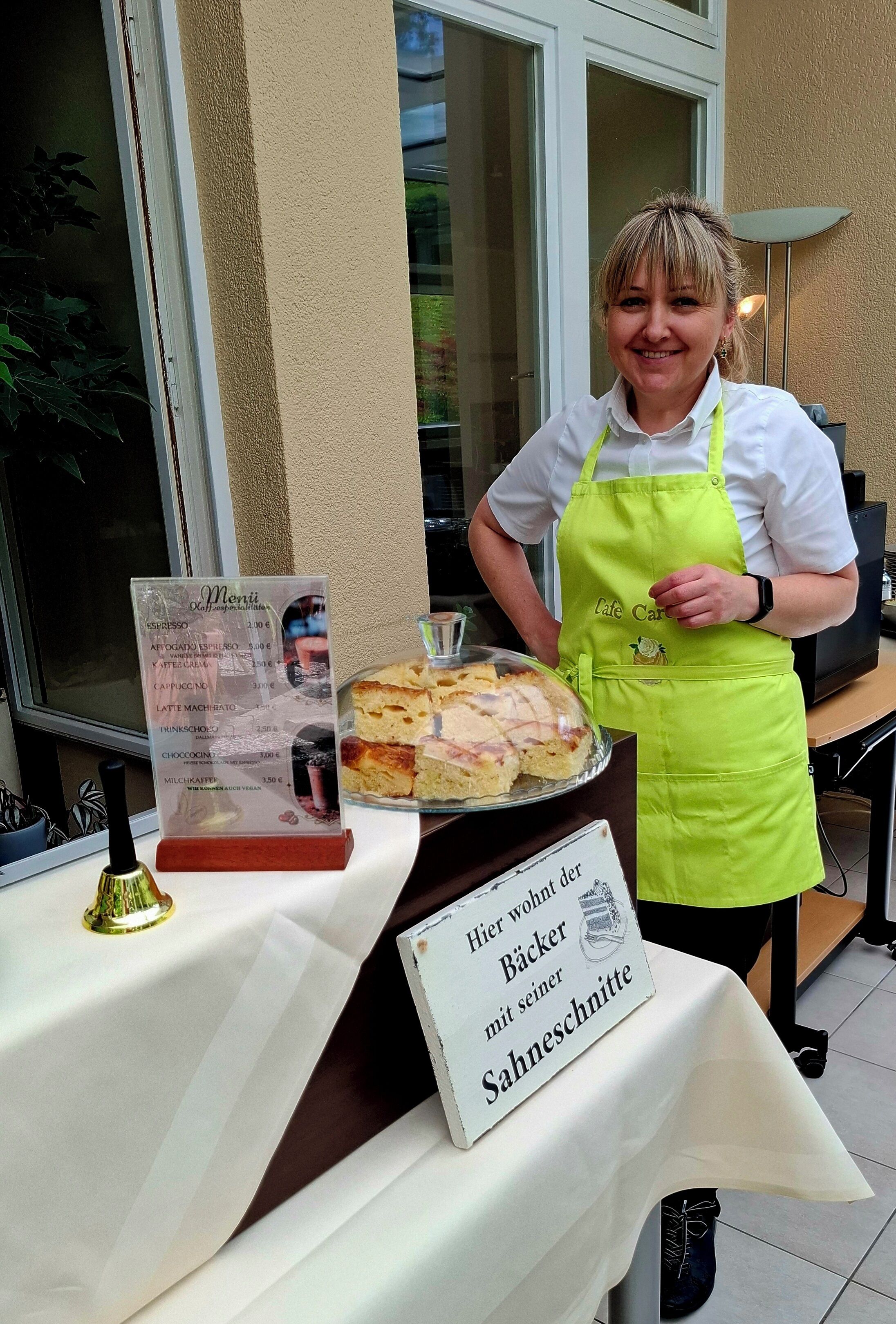 Une personne vêtue d’une chemise blanche et d’un tablier vert se tient derrière une table sur laquelle est posée une pancarte indiquant : « C’est ici que vivent les boulangers avec leur spécialité à la crème. »