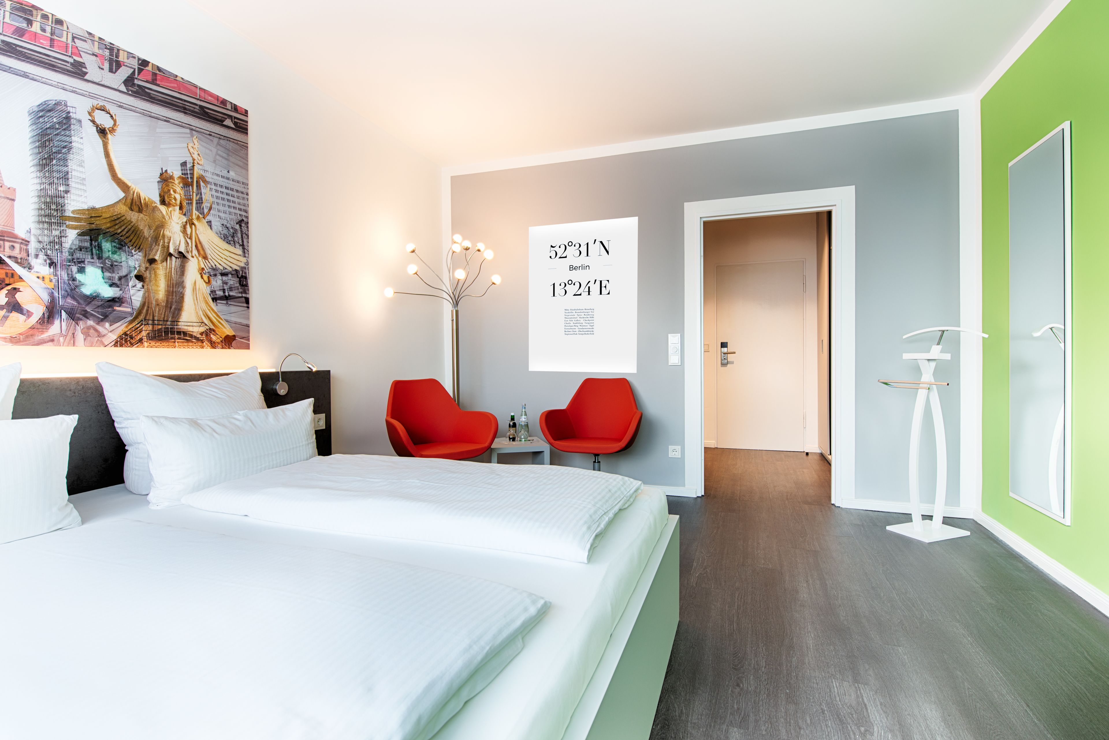 Hotellihuone, jossa on suuri sänky, kaksi punaista tuolia, pieni pöytä pulloineen, koordinaatteja esittävä juliste ja vihreä seinä.