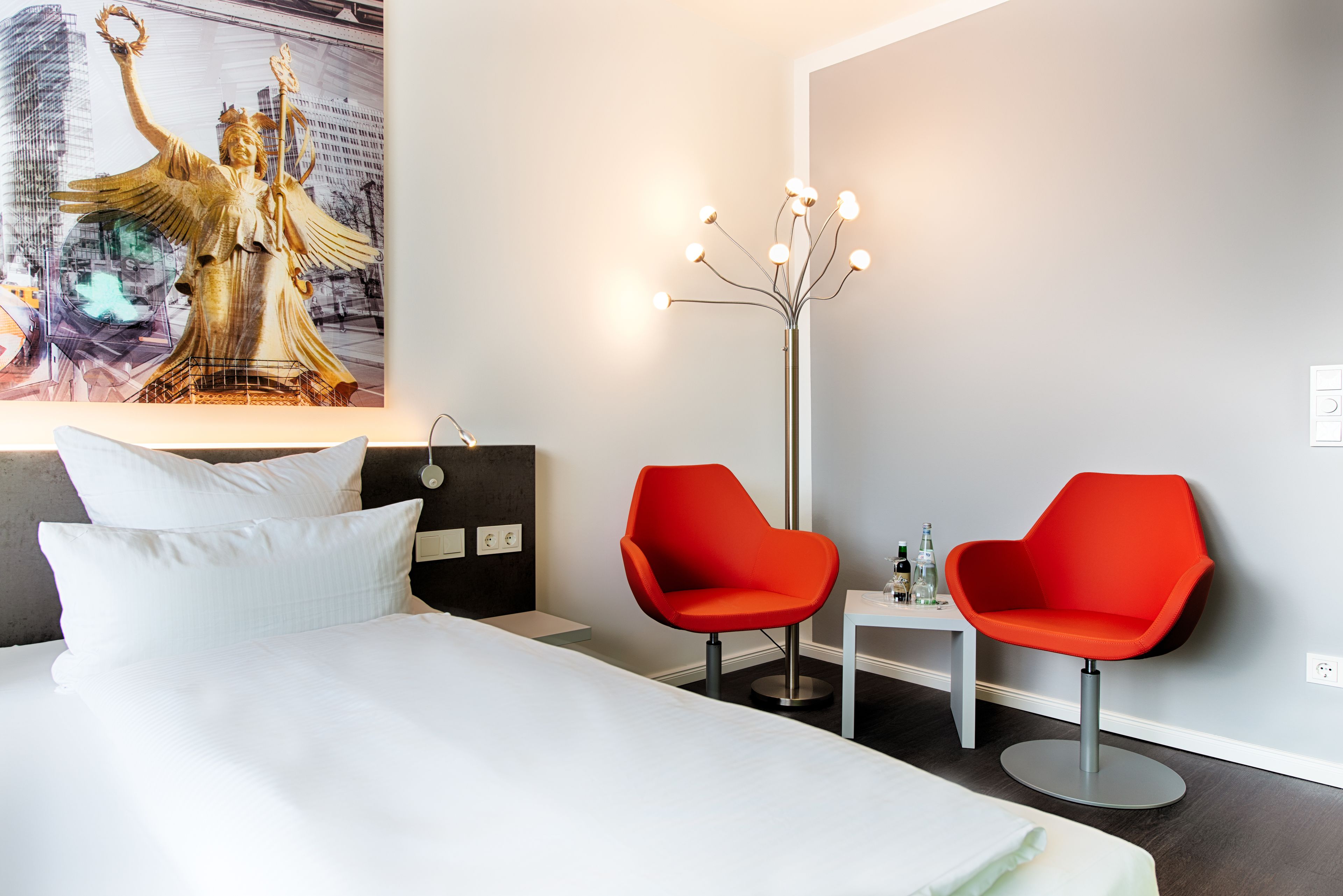 A hotel room with a bed, two red chairs, a small table, a floor lamp, and a painting of a statue.