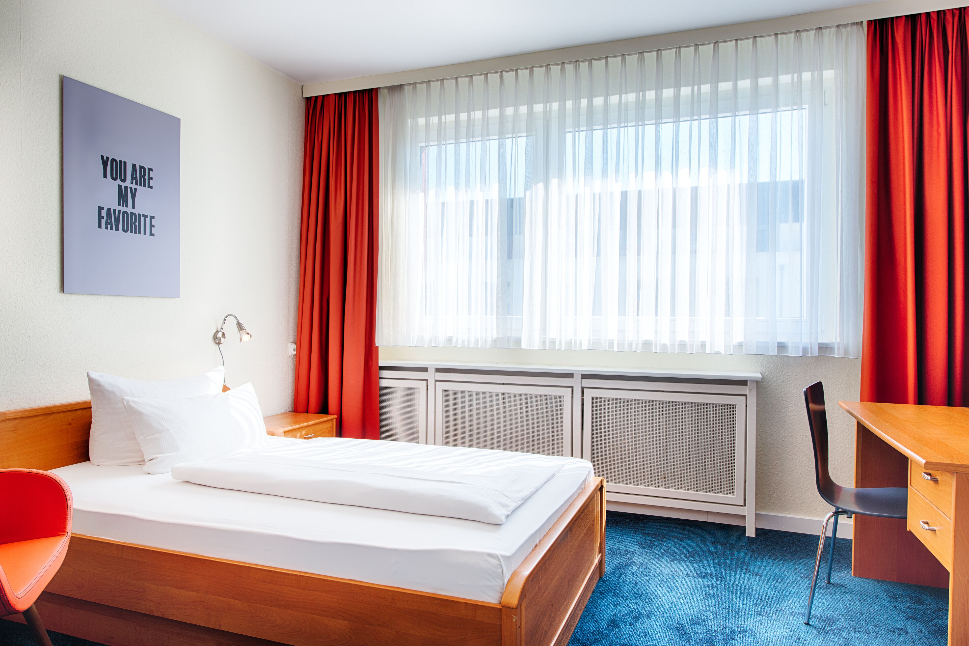 Una habitación de hotel con cama, escritorio, silla, radiador y ventana con cortinas.
