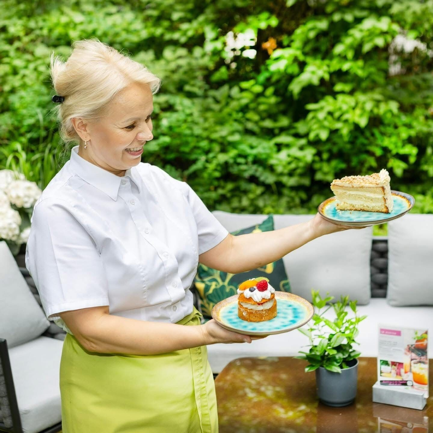 Una persona tiene in mano due piatti con dolci: uno con una fetta di torta e l'altro con una pasta sfoglia guarnita con panna e frutta, in un giardino con piante e un divano.