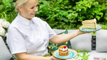 Una persona tiene in mano due piatti con dolci: uno con una fetta di torta e l'altro con una pasta sfoglia guarnita con panna e frutta, in un giardino con piante e un divano.