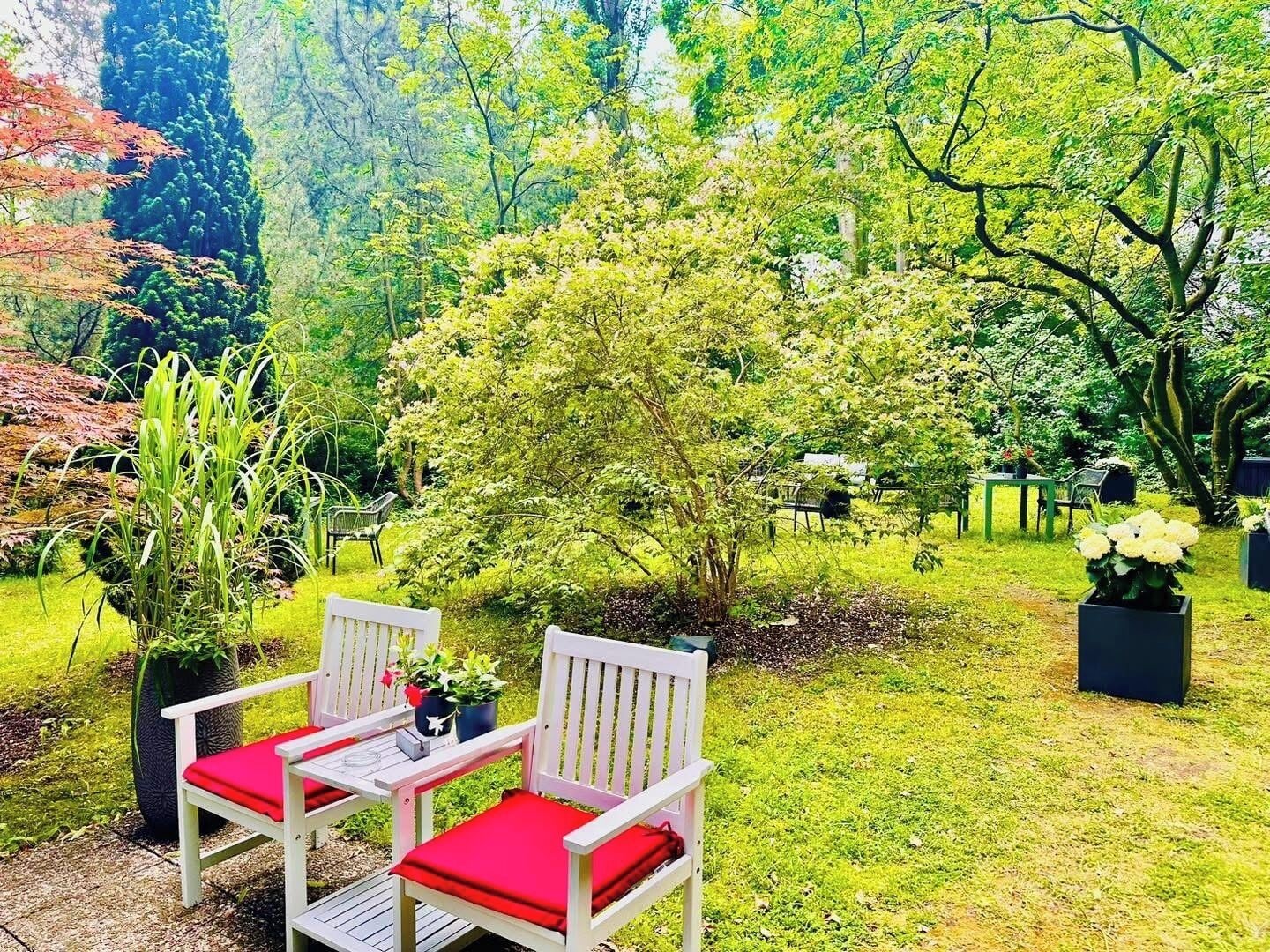 Un jardin avec deux chaises blanches dotées de coussins rouges, une petite table, ainsi que diverses plantes et arbres.