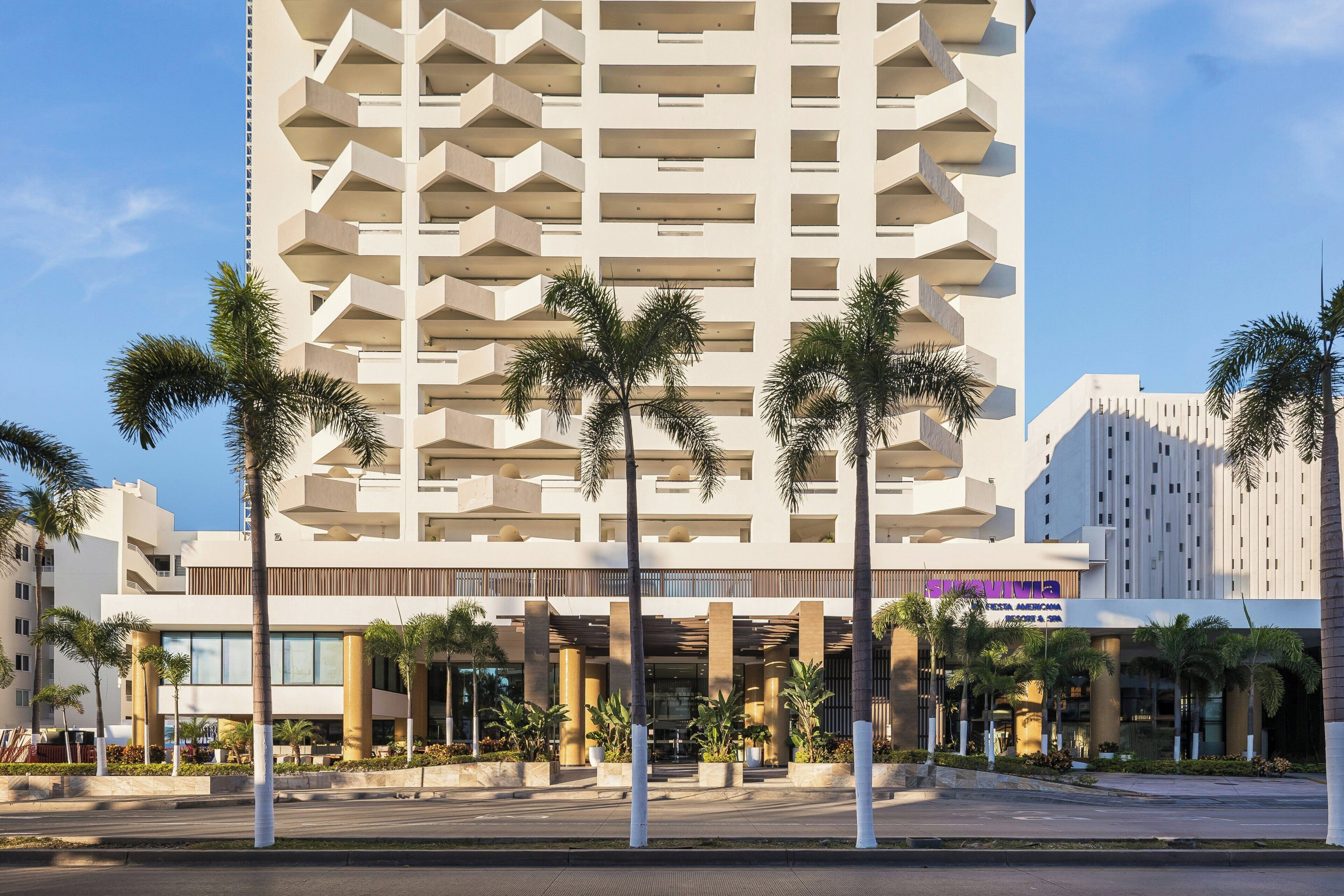 Ein modernes Hotel mit einer einzigartigen geometrischen Fassade, Palmen im Vordergrund und einem wolkenlosen Himmel.