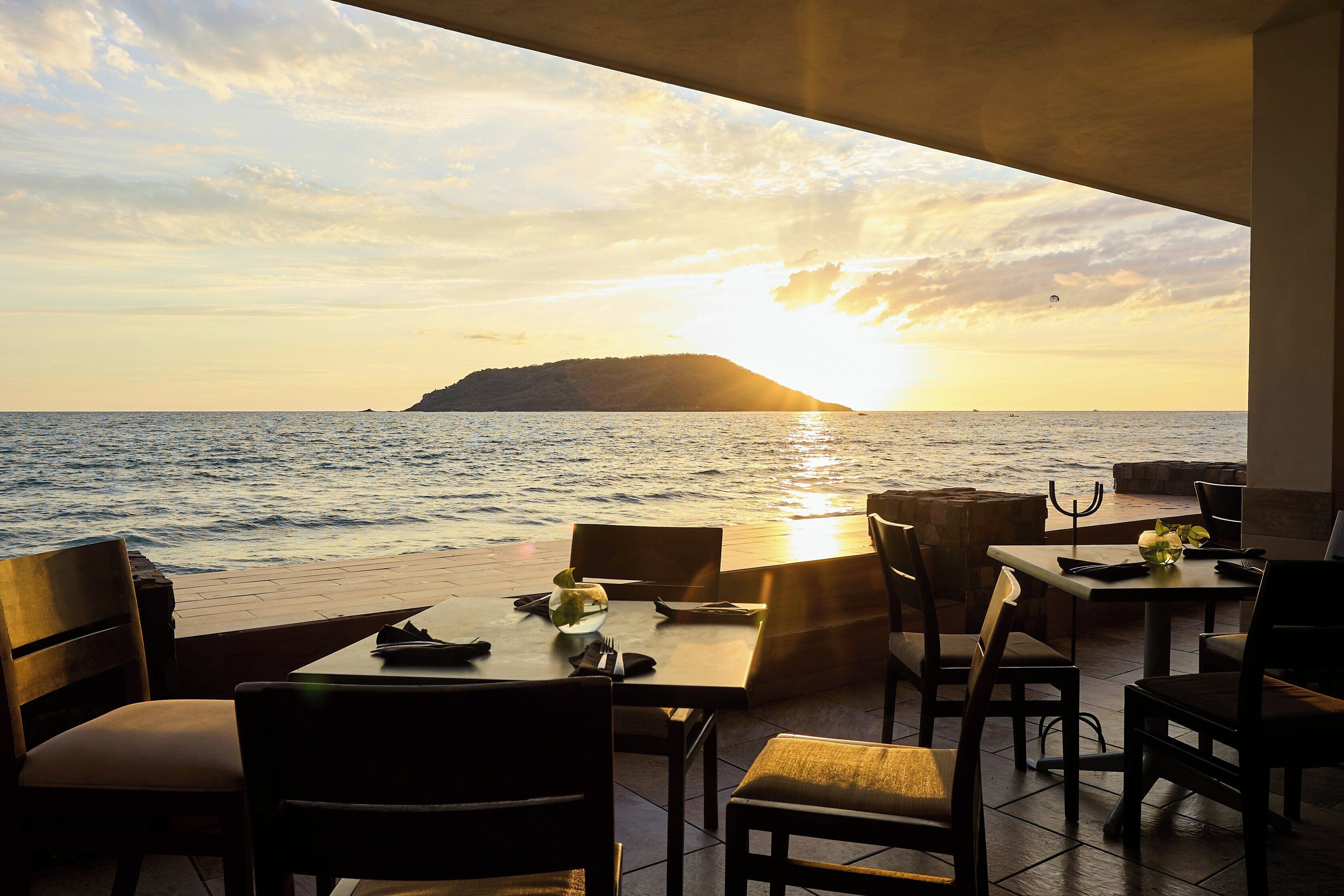 An outdoor dining area with tables and chairs facing a sunset over the sea.