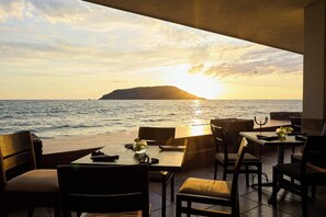 Un área de comedor al aire libre con mesas y sillas frente a un atardecer sobre el mar.