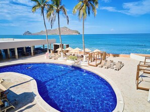 A swimming pool with lounge chairs and umbrellas, a beach view, and a mountainous backdrop.