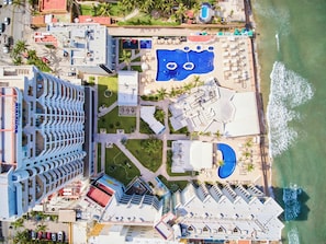Vista aérea de un complejo turístico con múltiples piscinas, una zona de playa y un estacionamiento.