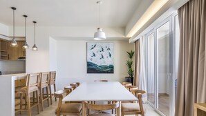 A modern dining area with a long table, wooden chairs, pendant lighting, and a large window with curtains.