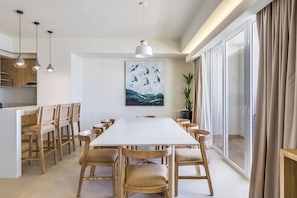 Un espace de salle à manger moderne, avec une longue table, des chaises en bois, des luminaires suspendus et une grande fenêtre agrémentée de rideaux.