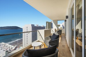A balcony with wicker chairs and a small table, overlooking a beach and cityscape.