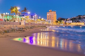 Una playa con costa rocosa, palmeras y un edificio de varios pisos.