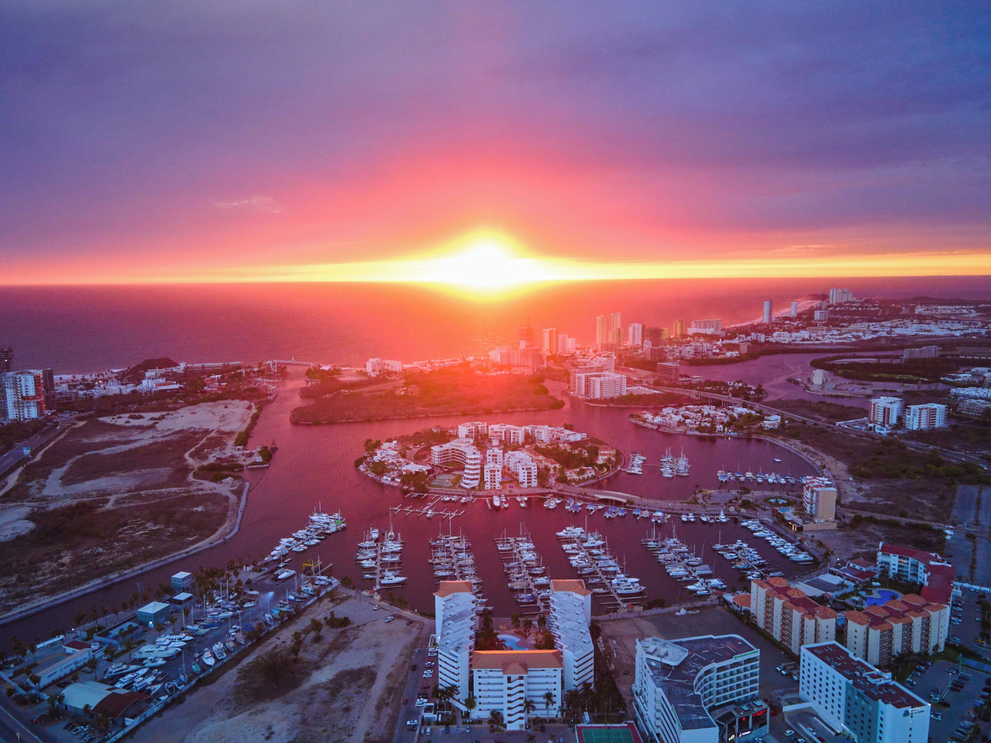 An aerial view of a marina with boats docked, surrounded by buildings and a sunset in the background.