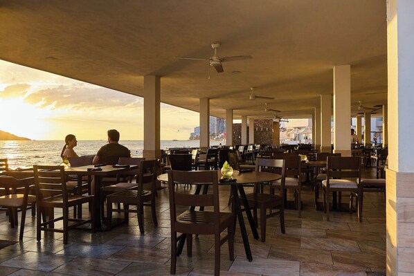 Un espacio de comedor al aire libre con mesas y sillas, con vista a un cuerpo de agua y un paisaje urbano de fondo.