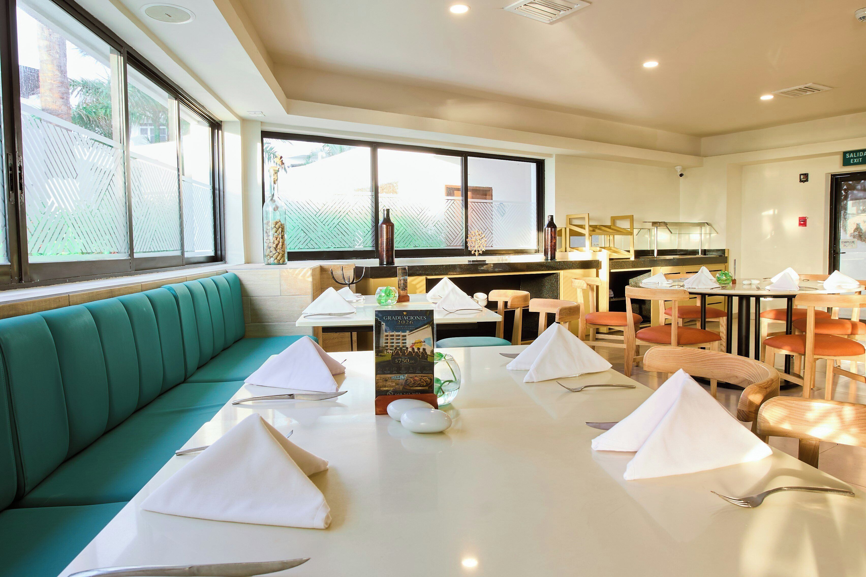 A restaurant interior with a long table set for dining, a green cushioned bench, and a window with a view.