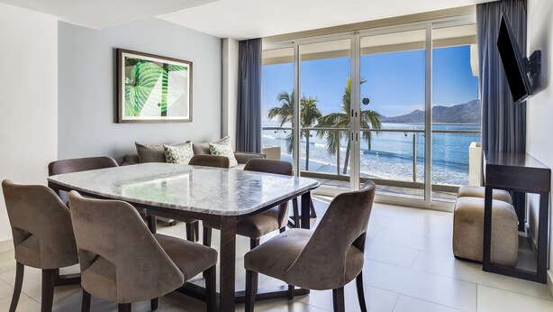 A modern dining area with a large table and chairs, a flat-screen TV mounted on the wall, and a large window offering a view of the sea and mountains.