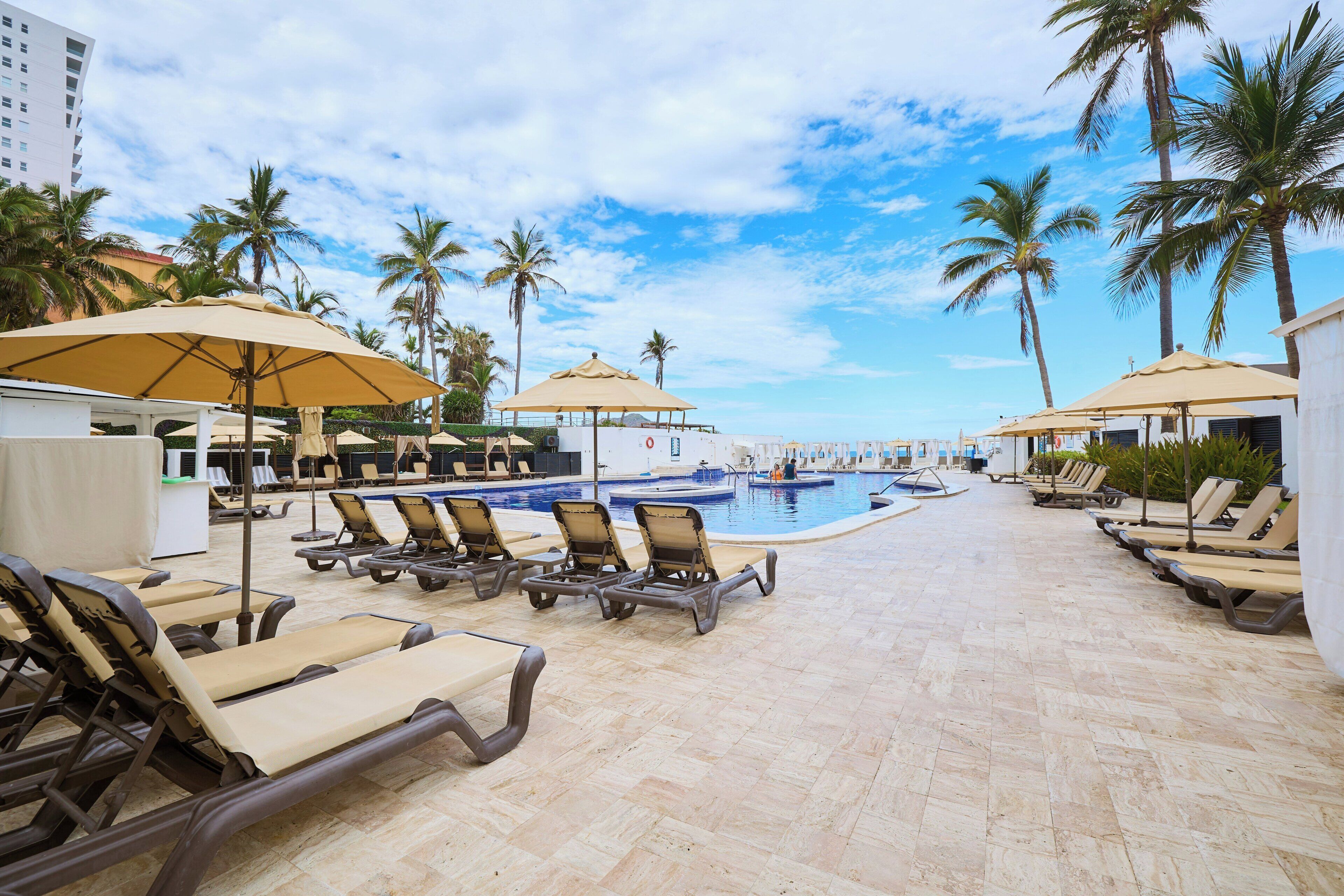 A poolside area with lounge chairs and umbrellas, palm trees, and a clear sky.