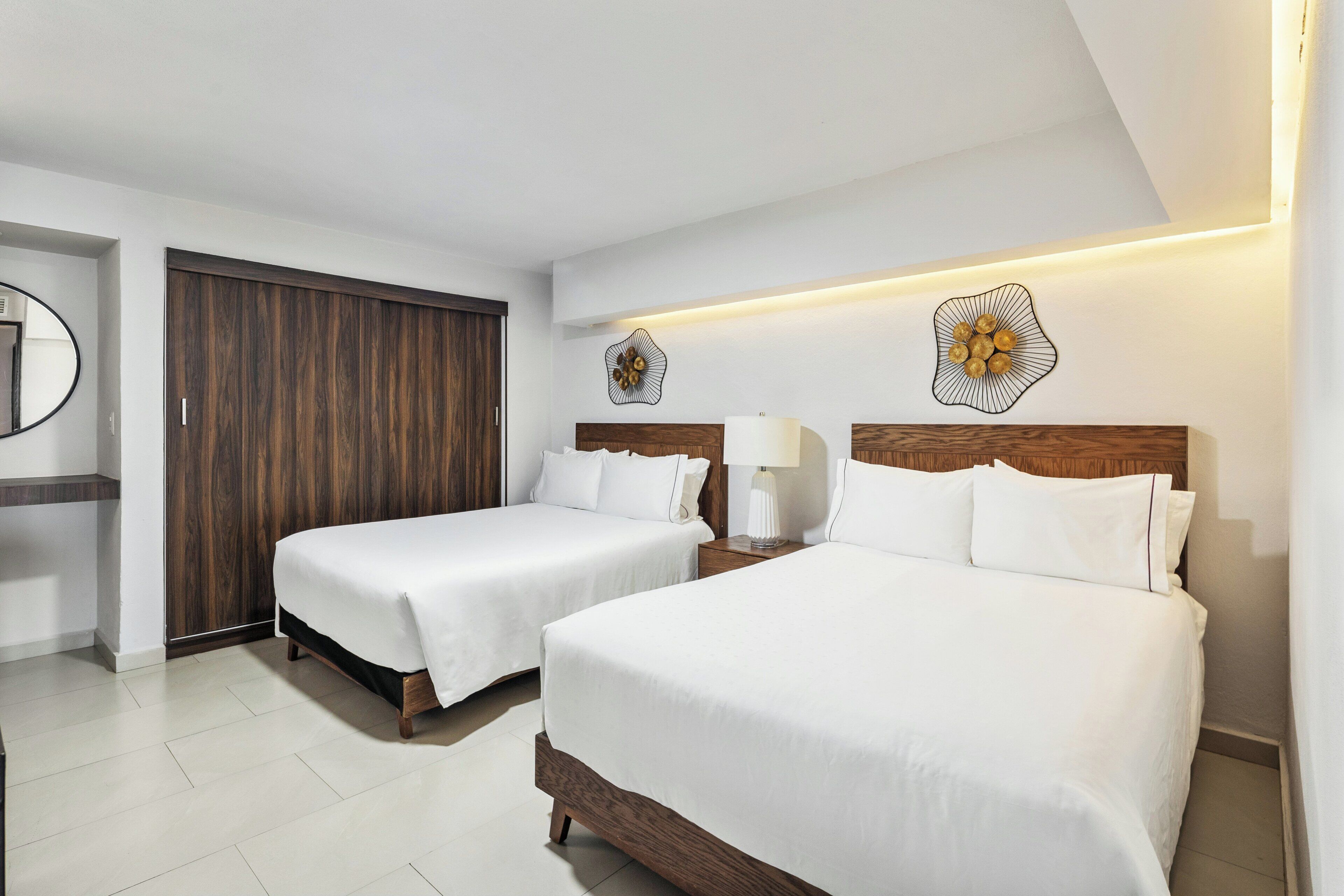 A hotel room with two beds, a wooden wardrobe, a mirror, and decorative wall art.