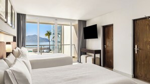 A hotel room with two beds, a TV, a desk, and a balcony with a view of the beach and mountains.