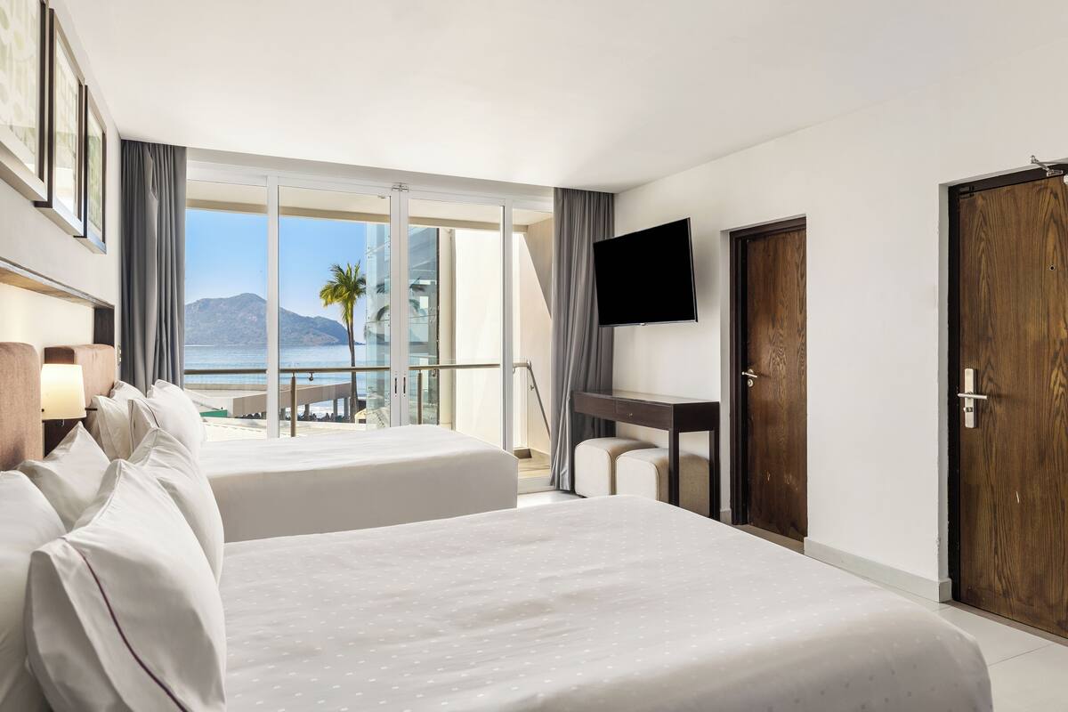 Une chambre d’hôtel avec deux lits, une télévision, un bureau et un balcon offrant une vue sur la plage et les montagnes.