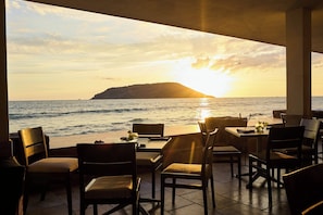 Un comedor con mesas y sillas de frente a una puesta de sol sobre el mar.
