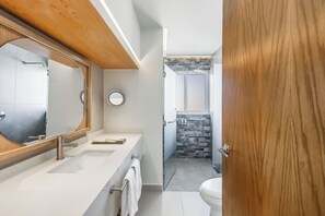 Ein modernes Badezimmer mit Holzboden, einem großen Spiegel, zwei Waschbecken, einer Toilette und einer Duschkabine mit Steinwand.