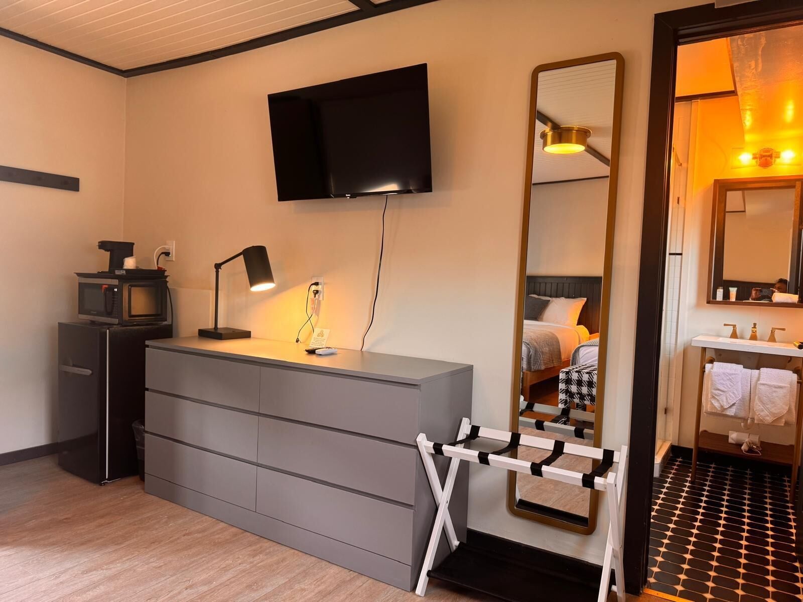 Habitación de hotel con televisión de pantalla plana, escritorio con lámpara, espejo, cama y baño con ducha.