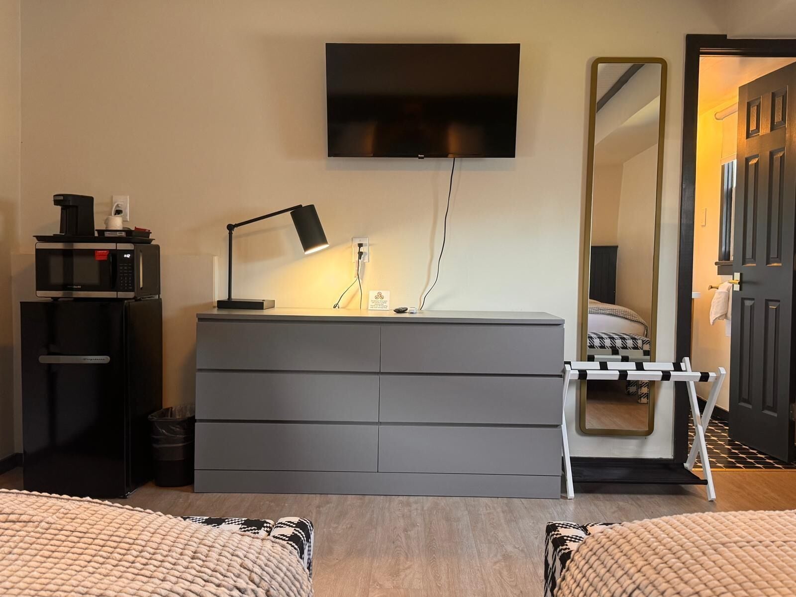 Une chambre d’hôtel avec une commode grise, un téléviseur à écran plat fixé au mur, une table de chevet avec une lampe, un lit recouvert d’une couvre-lit à motifs et un miroir sur le mur.