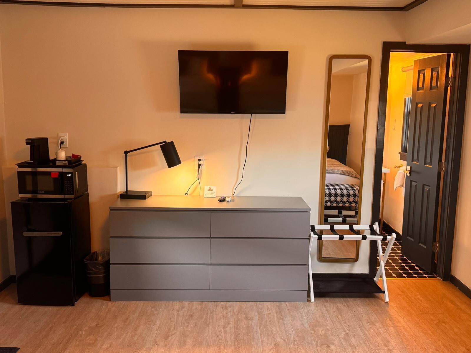 A hotel room with a grey dresser, a flat-screen TV mounted above it, a bedside table with a lamp, a trash bin, and a mirror reflecting the room.