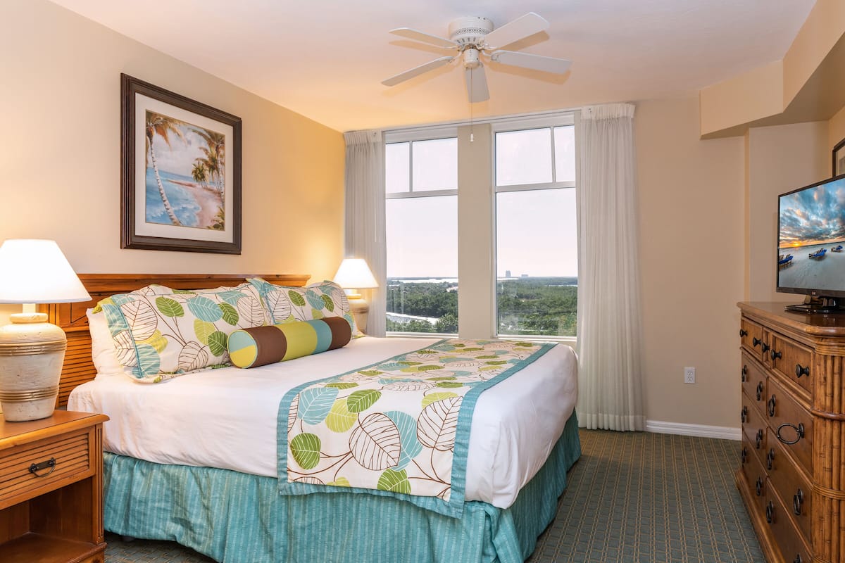 A hotel room with a bed, a television, a ceiling fan, a window with curtains, and a framed picture on the wall.