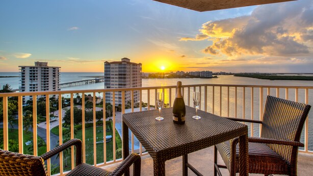 A balcony with wicker furniture, a bottle, and glasses, overlooking a sunset over a body of water and buildings. - Lovers Key Resort (Fort Myers Beach)