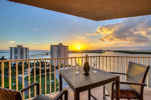 A balcony with wicker furniture, a bottle, and glasses, overlooking a sunset over a body of water and buildings. - Lovers Key Resort (Fort Myers Beach)